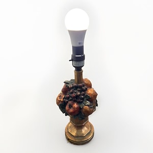 May include: A table lamp with a fruit-themed base. The base is gold-coloured and features a detailed arrangement of sculpted fruits, including grapes, apples, and pears. The lamp has a black socket and a bright white LED light bulb.
