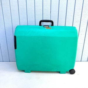 May include: A bright teal hard-shell suitcase featuring a black top handle, a gold-toned locking mechanism, and a single visible black wheel at the bottom right corner. The rectangular luggage has a smooth, solid-colored exterior surface.
