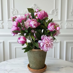 May include: A bouquet of pink and white peonies in a weathered green ceramic pot with a terracotta saucer. The flowers have multiple layers of petals and green leaves. The arrangement sits on a white marble table.