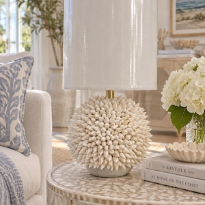 May include: A table lamp featuring a spherical base covered in numerous small, off-white, pointed sea shells. The lamp has a tall, cylindrical, smooth white shade and a small gold-toned metal neck connecting the base to the shade.