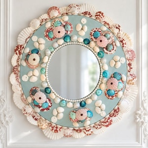 May include: A round wall mirror featuring a light blue frame decorated with an assortment of white, brown, and teal seashells. The outer edge is lined with white scallop shells, while the inner border is accented with small white beads.