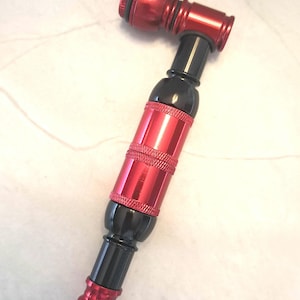 May include: A red and black metal pipe with a curved design. The pipe features a cylindrical body with textured bands and a rounded mouthpiece. The stem is black, connecting the red components. The pipe is designed for smoking.