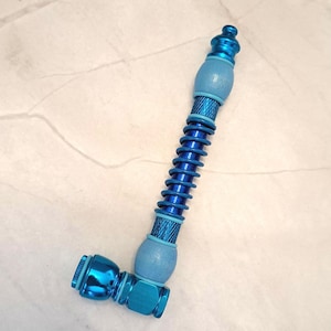 May include: A blue metal pipe with a curved design. The pipe features a spiral section, two rounded sections, and a bowl at the end. The pipe is a vibrant shade of blue and has a shiny finish.
