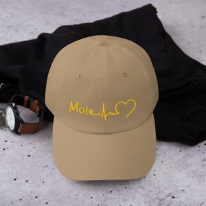 Könnte beinhalten: Beige Baseballkappe mit dem Wort "Moin" und einer gelben Herzschlaglinie, die in einem Herzen endet. Die Kappe ist aus weichem Material und hat einen gebogenen Schirm. Ein lässiges Accessoire.