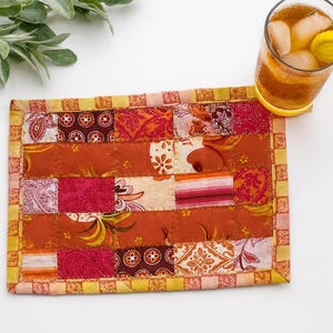 May include: A rectangular, patchwork coaster with a variety of orange, red, and cream-colored fabrics. The coaster has a yellow border and is next to a glass of iced tea with a lemon slice.