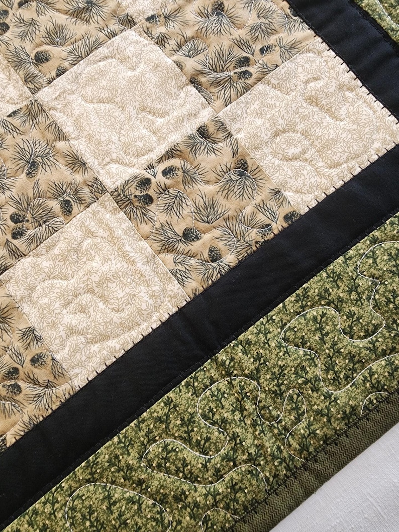 K&ouml;nnte beinhalten: Nahaufnahme eines gesteppten Stoffes mit einem von der Natur inspirierten Design. Der Quilt zeigt quadratische Bl&ouml;cke mit beigem Hintergrund und einem Muster aus Kiefernadeln und Tannenzapfen in Dunkelgrau. Ein schwarzer Rand trennt die gemusterten Abschnitte von einem gr&uuml;nen, blattf&ouml;rmigen Rand.