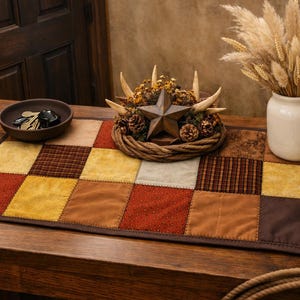 May include: A rustic table runner with a patchwork design in shades of brown, gold, and red. A decorative star and antler wreath sits atop the runner, along with a vase of dried flowers and a small bowl of keys.