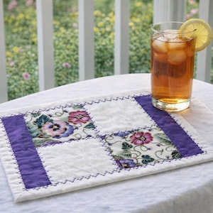 May include: A quilted placemat with a floral design and purple accents sits on a white tablecloth. A glass of iced tea with a lemon slice rests on the placemat. The background features a white railing and greenery.