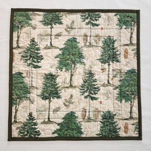 K&ouml;nnte beinhalten: Quadratische, gesteppte Tischdecke mit einem sich wiederholenden Muster aus gr&uuml;nen B&auml;umen und Tannenzapfen auf cremefarbenem Hintergrund. Die Tischdecke hat einen dunkelgr&uuml;nen Rand. Die Steppung verleiht Textur. Wahrscheinlich aus Baumwolle oder &auml;hnlichem Stoff.