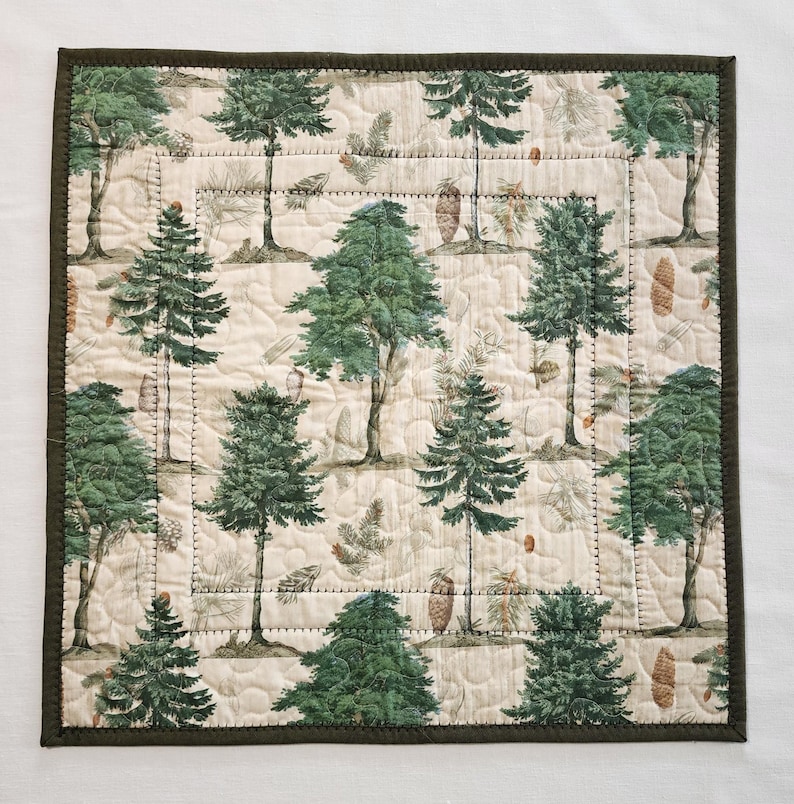 K&ouml;nnte beinhalten: Quadratische, gesteppte Tischdecke mit einem sich wiederholenden Muster aus gr&uuml;nen B&auml;umen und Tannenzapfen auf cremefarbenem Hintergrund. Die Tischdecke hat einen dunkelgr&uuml;nen Rand. Die Steppung verleiht Textur. Wahrscheinlich aus Baumwolle oder &auml;hnlichem Stoff.