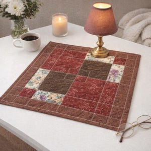 May include: A square, quilted placemat with a patchwork design in shades of brown, burgundy, and floral patterns. A small lamp with a burgundy shade sits on the placemat, along with a cup of coffee and a lit candle.