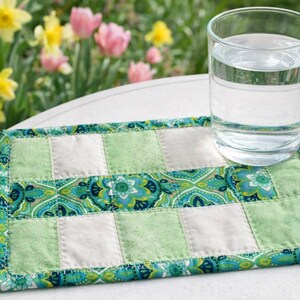 May include: A clear glass of water sits on a handmade, quilted coaster. The coaster features a patchwork design with green, white, and teal paisley fabric. The background includes a garden with pink and yellow flowers.