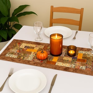 May include: A table setting with a brown patchwork table runner, white plates, and lit candles. A small orange pumpkin sits on the runner. Two wooden chairs are in the background. A large green plant is in a white pot.