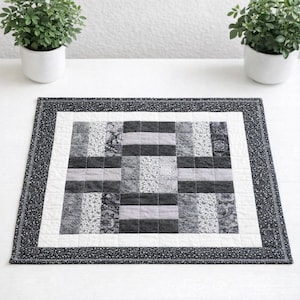 May include: A rectangular quilted placemat with a patchwork design in shades of gray and white. The center features a grid pattern, surrounded by a white border and a black floral patterned edge. The placemat is likely handmade.