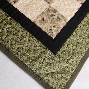 K&ouml;nnte beinhalten: Nahaufnahme eines gesteppten Tischl&auml;ufers mit Patchwork-Design. Der L&auml;ufer zeigt Quadrate aus beigem Stoff mit Tannenzapfenmustern, die von schwarzem Stoff und einem gr&uuml;nen, blattartigen Druck umrandet sind. Die Kanten sind mit einer dunkelgr&uuml;nen Paspel versehen.