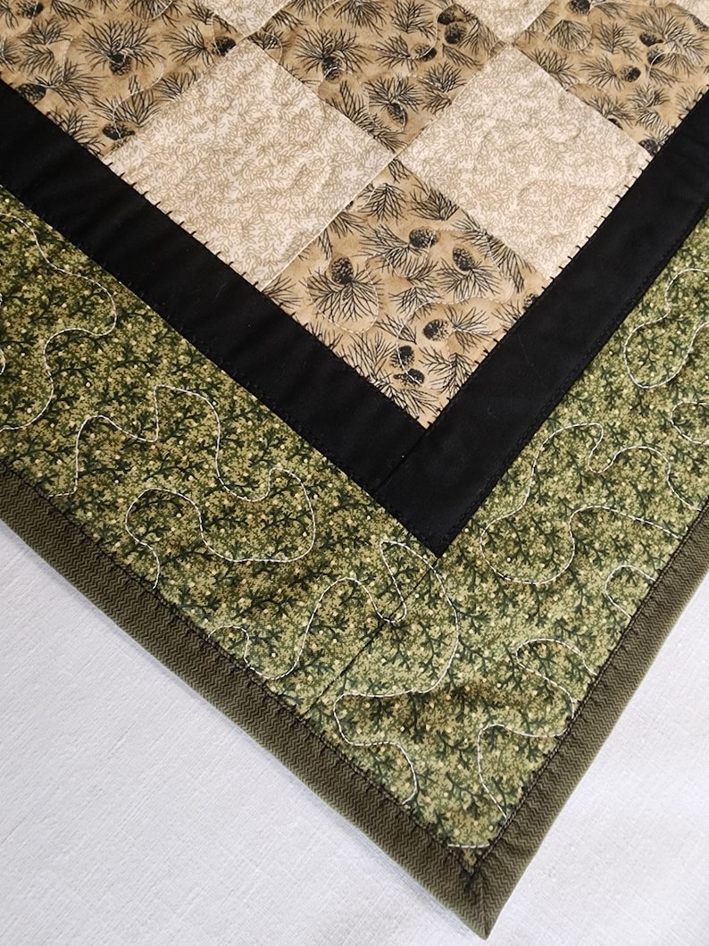 K&ouml;nnte beinhalten: Nahaufnahme eines gesteppten Tischl&auml;ufers mit Patchwork-Design. Der L&auml;ufer zeigt Quadrate aus beigem Stoff mit Tannenzapfenmustern, die von schwarzem Stoff und einem gr&uuml;nen, blattartigen Druck umrandet sind. Die Kanten sind mit einer dunkelgr&uuml;nen Paspel versehen.