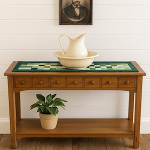 May include: A wooden console table with a green and white patterned runner, a cream-colored pitcher and basin, and a framed portrait. A potted plant sits on the lower shelf. The table has seven small drawers.