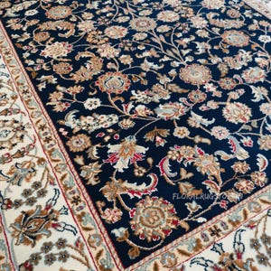 May include: A navy blue floral area rug with a cream border. The rug features a detailed pattern of flowers and leaves in shades of beige, brown, red, and blue. The rug is rectangular and appears to be made of a soft material.