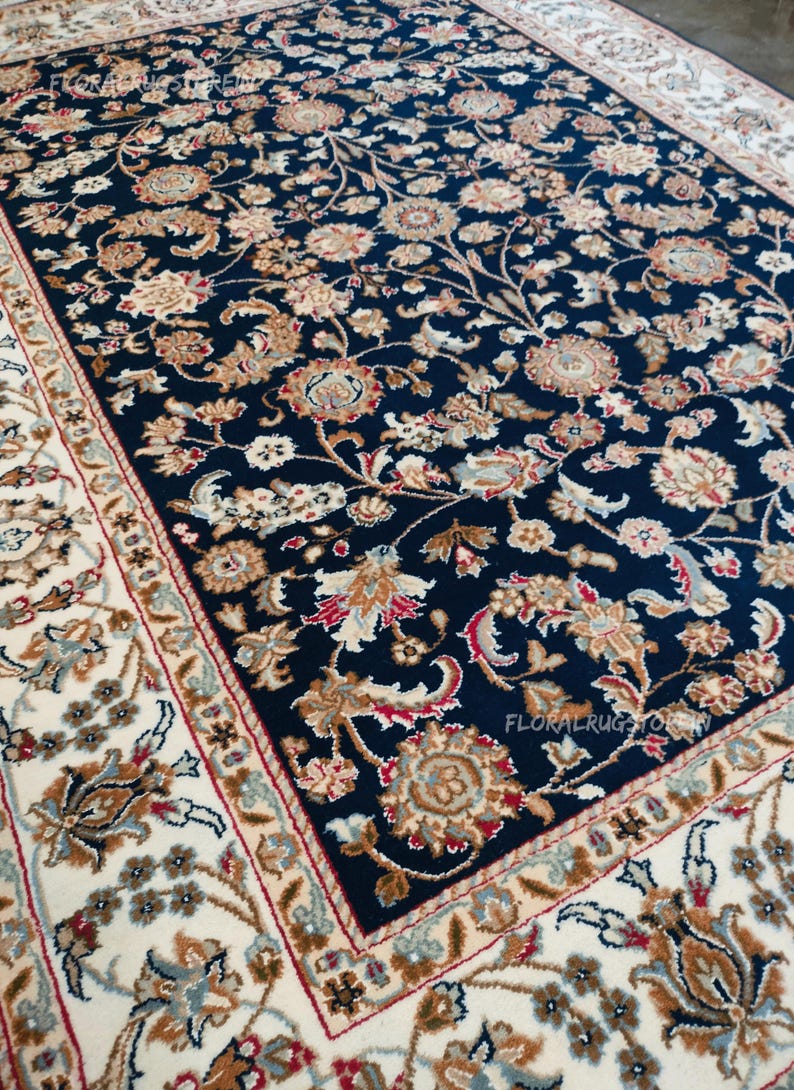 May include: A navy blue floral area rug with a cream border. The rug features a detailed pattern of flowers and leaves in shades of beige, brown, red, and blue. The rug is rectangular and appears to be made of a soft material.