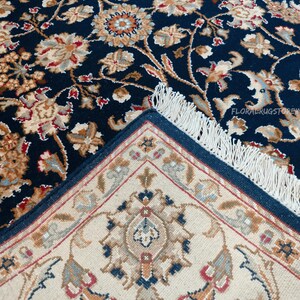 May include: A detailed shot of a navy blue rug with a floral pattern in beige, brown, and red. The rug's edge reveals a cream-colored underside with a geometric design and a navy blue border. The rug has a fringe.