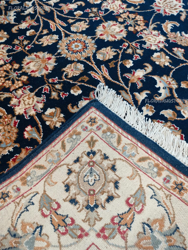 May include: A detailed shot of a navy blue rug with a floral pattern in beige, brown, and red. The rug's edge reveals a cream-colored underside with a geometric design and a navy blue border. The rug has a fringe.