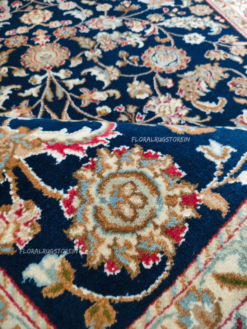 May include: Close-up of a navy blue rug with an intricate floral pattern in shades of beige, red, and light blue. The rug features a detailed design with scrolling vines and stylized flowers. The text "FLORALRUGSTOREIN" is visible.