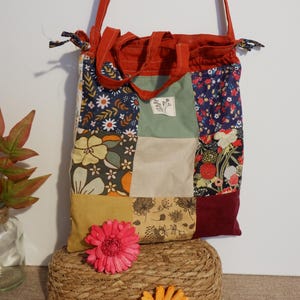 Op de afbeelding: Een patchwork tote bag met een rode band en trekkoordsluiting. De tas heeft een verscheidenheid aan bloemmotieven in blauw, geel, groen en rood. Een roze en oranje bloem ligt voor de tas.