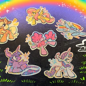 May include: A collection of colorful cartoon pony stickers with glitter accents. The stickers feature various ponies in different poses, set against a background with a rainbow, clouds, and stars.