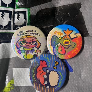 May include: Three circular pin-back buttons with a holographic finish. One button features a character with the text AH! WHAT A PLEASANT AROMA!. Another shows a colourful creature with the text BLEP. The third button depicts a shrimp-like figure.
