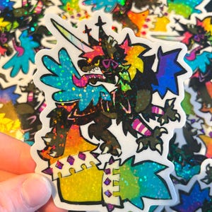 May include: A colorful sticker featuring a fantasy creature with a unicorn horn, wings, and a spiky tail. The sticker has a glittery, holographic finish with shades of blue, green, yellow, and pink. The creature has a fierce expression.
