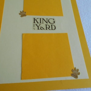 May include: A white background with a yellow border and two yellow squares. The top square has the text "KING of the YARD" in brown lettering. The bottom square is blank. There are two paw print stickers on the image.