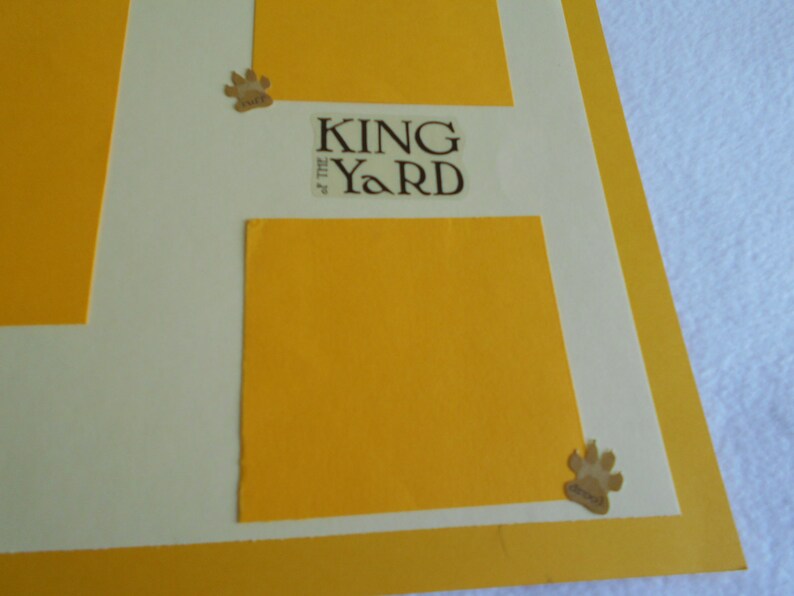 May include: A white background with a yellow border and two yellow squares. The top square has the text "KING of the YARD" in brown lettering. The bottom square is blank. There are two paw print stickers on the image.