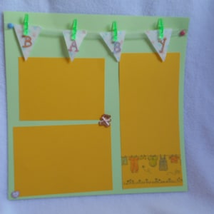May include: A green cardstock scrapbook page with yellow photo mats and a banner that says "BABY" in brown letters. The banner is made of white fabric with a floral pattern and is strung with green clothespins. There are also some small decorative items on the page, including a brown teddy bear.