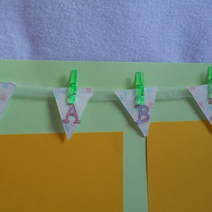 May include: A green and white baby banner with the word "BABY" spelled out in pink and orange letters. The banner is hanging from green clothespins on a light green background.