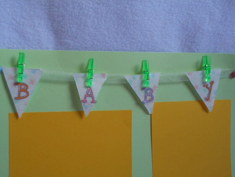 May include: A green and white baby banner with the word "BABY" spelled out in pink and orange letters. The banner is hanging from green clothespins on a light green background.