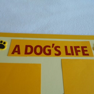 May include: A yellow and cream-colored scrapbook with a red title that reads "A Dog's Life". The title is surrounded by two black paw prints with yellow outlines.