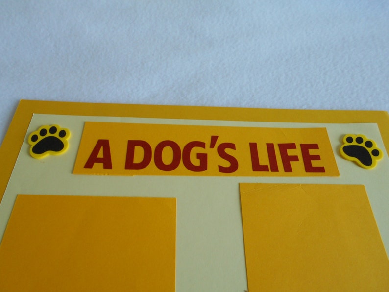 May include: A yellow and cream-colored scrapbook with a red title that reads "A Dog's Life". The title is surrounded by two black paw prints with yellow outlines.