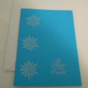 Tarjeta de Navidad hecha a mano, los copos de nieve con paz en la tierra estampado en blanco en la parte delantera de la tarjeta azul, con juego sobre blanco