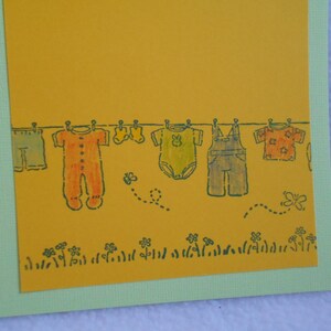 May include: A yellow card with a line of clothes hanging on a clothesline. The clothes include a pair of overalls, a long-sleeved onesie, a short-sleeved onesie, a pair of pants, and a short-sleeved shirt. There are also two butterflies and a bee on the card.