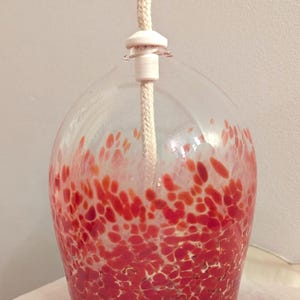 May include: A clear glass oil lamp with a red speckled pattern. The lamp has a rounded shape and a white wick. The wick extends from the top of the lamp. The lamp is designed for decorative purposes.