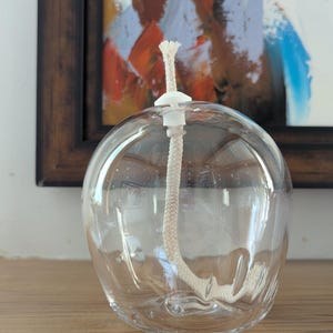 May include: A clear glass oil lamp with a spherical body and a white wick holder. The wick is a beige rope. The lamp is on a wooden surface, with a framed painting behind it.