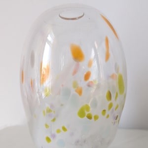 May include: A clear glass vase with a rounded form and a small opening at the top. The vase features colourful speckled patterns in orange, yellow, green, and purple. The vase is on a white surface.