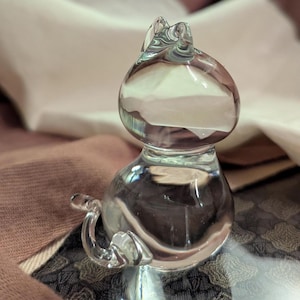 May include: A clear glass cat figurine with a rounded body and head, and a curled tail. The cat's ears are pointed, and the glass has a slight green tint. The figurine is sitting on a patterned surface.