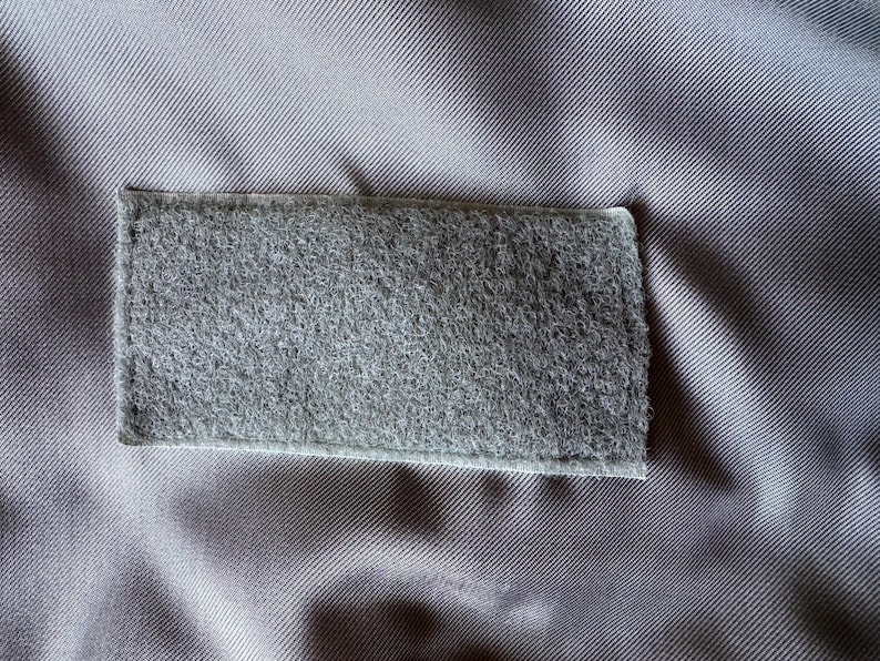 Op de afbeelding: Een rechthoekige grijze klittenbandpatch genaaid op een gladde, lichtgrijze stoffen achtergrond. De patch heeft een pluizig, getextureerd oppervlak dat kenmerkend is voor de lus-kant van een sluiting, met zichtbare stiksels langs de randen.