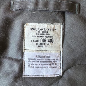 Op de afbeelding: De grijze gewatteerde binnenkant van een jas met een rechthoekig wit label met zwarte tekst: JACKET, FLYER'S, CWU 45/P, MIL-J-83388 D, DLA100-86-C-0353, 100% AROMATIC POLYAMIDE, X-LARGE (46-48), 8415-00-310-1140, AVIREX LTD. INSTRUCTION LABEL.