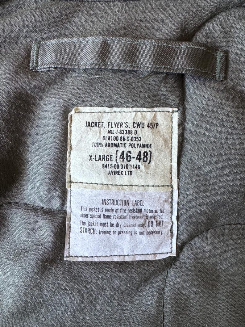 Op de afbeelding: De grijze gewatteerde binnenkant van een jas met een rechthoekig wit label met zwarte tekst: JACKET, FLYER'S, CWU 45/P, MIL-J-83388 D, DLA100-86-C-0353, 100% AROMATIC POLYAMIDE, X-LARGE (46-48), 8415-00-310-1140, AVIREX LTD. INSTRUCTION LABEL.