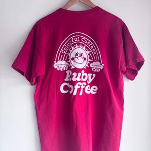 Op de afbeelding: Een magenta t-shirt met een witte afbeelding op de achterkant. De afbeelding toont een lachende zon in een regenboog, met de woorden "Colorful Coffees" en "Ruby Coffee" in een speels lettertype. Het shirt hangt aan een houten hanger.