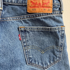 May include: Close-up of blue denim jeans with a back pocket and a brown leather Levi Strauss & Co. label. The label reads "505" and "W 30 32". The jeans have yellow stitching and a small red tag on the pocket.
