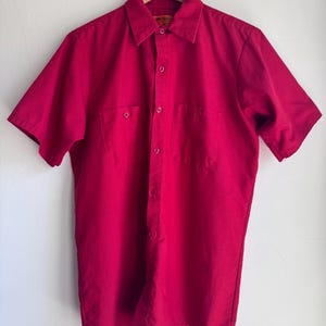 May include: A short-sleeved, button-down shirt in a vibrant red hue. The shirt features two chest pockets and a classic collar. It is hanging on a wooden hanger against a plain white background. The shirt appears to be made of a durable fabric.