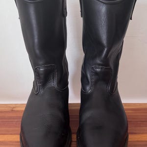 May include: A pair of black leather mid-calf boots featuring a rounded toe, a structured shaft with pull tabs at the top, and visible stitching details along the vamp. The boots have a smooth finish and a sturdy sole construction.